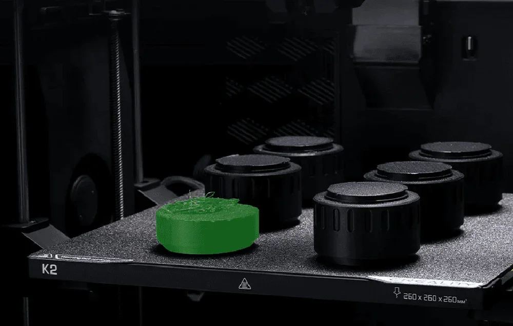 3D spausdintuvas su žaliu modeliu ant spausdinimo platformos šalia juodų rankenėlių