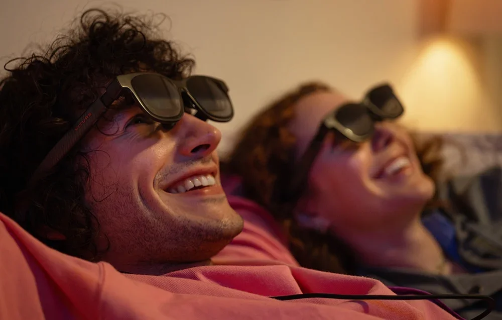 Moteris ir vyras su XR akiniais juokiasi gulėdami kambaryje su šiltu apšvietimu
