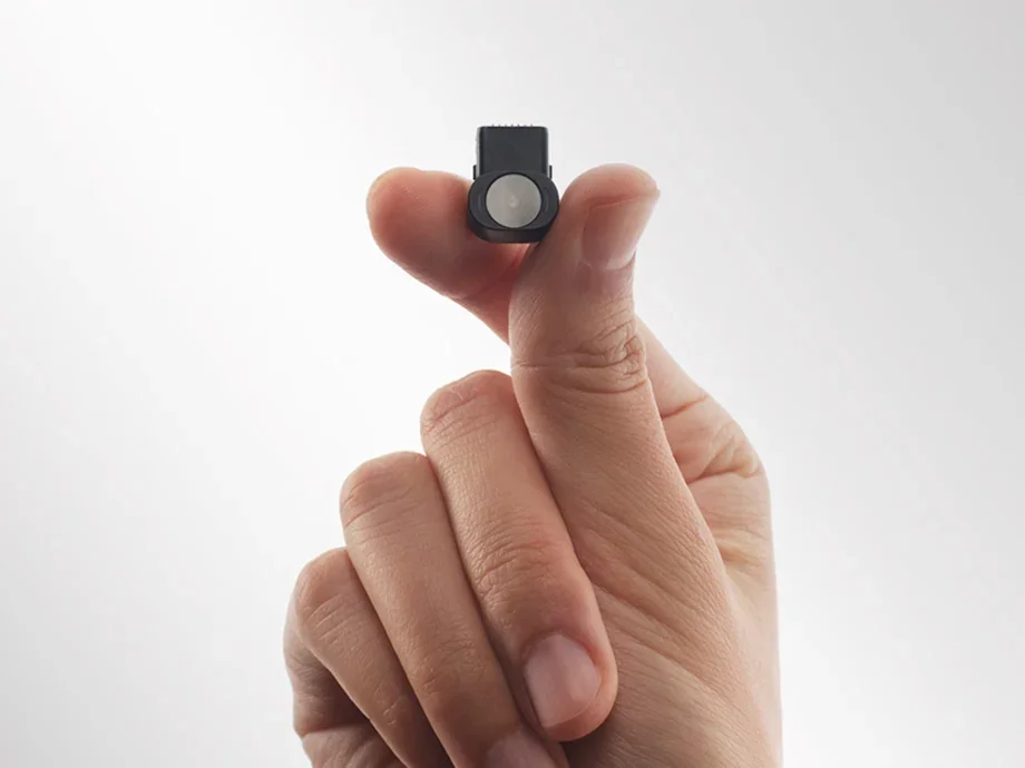 A small black camera held between the index finger and thumb against a bright background.