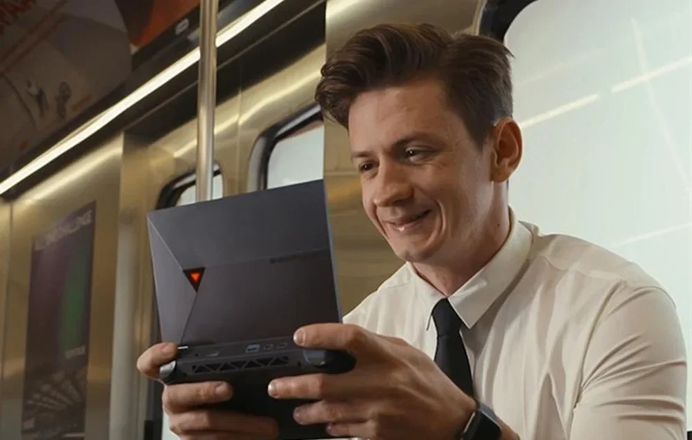 A man in a white shirt and tie plays a handheld console on a train, holding a gray device with controllers