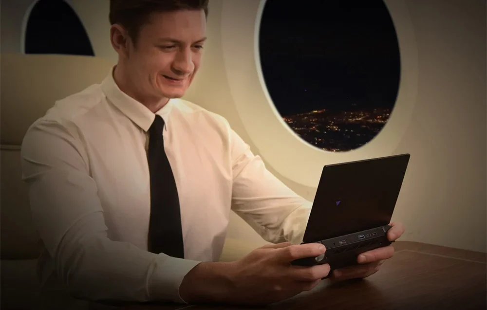 A man in a white shirt works on a small laptop on an airplane by a window overlooking the night city