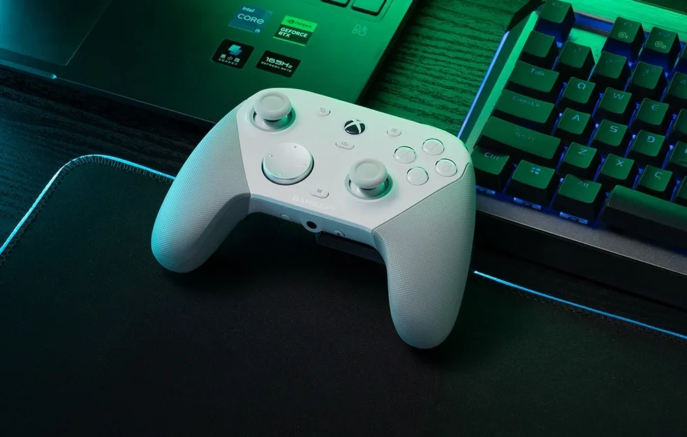Baltas GameSir valdiklis ant kilimėlio šalia nešiojamojo kompiuterio ir klaviatūros.
