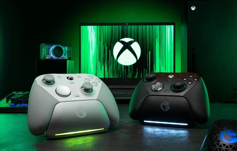 Du GameSir valdikliai įkrovimo stotelėje priešais monitorių su Xbox logotipu, vienas baltas, kitas juodas.