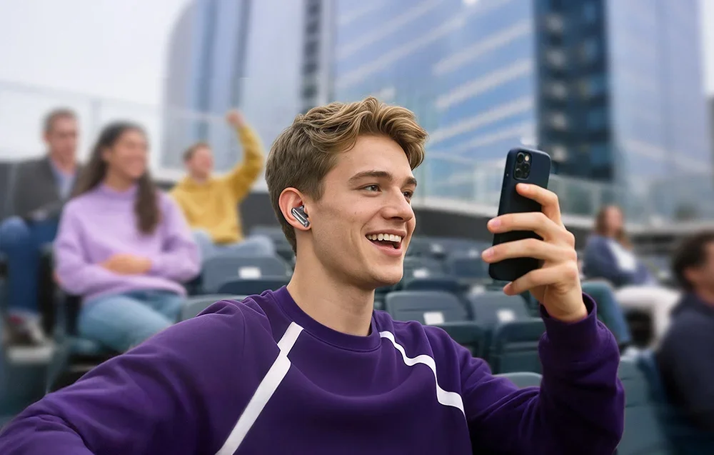 Jaunas vyras violetiniu džemperiu sėdi tribūnose, laiko išmanųjį telefoną ir naudoja ausinę